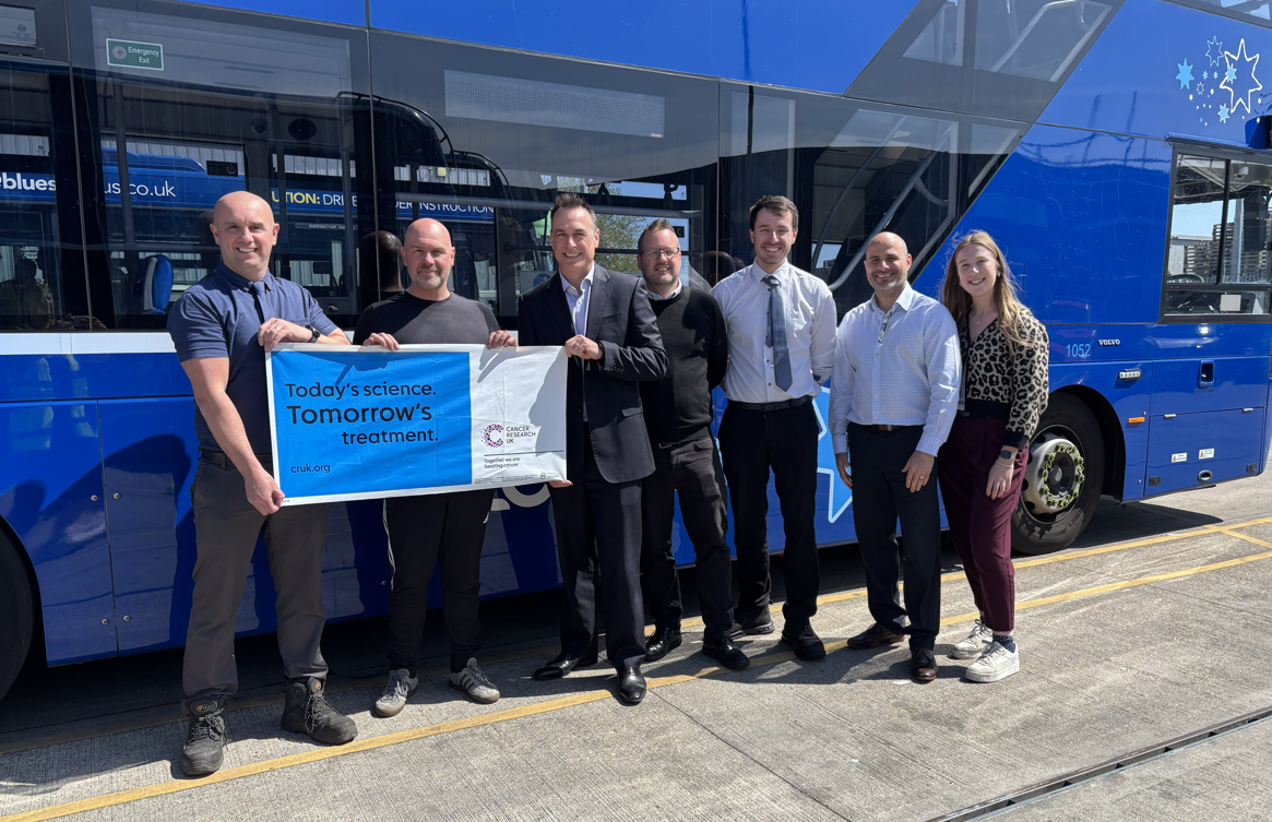Group of people stood next to a blue bus holding a banner