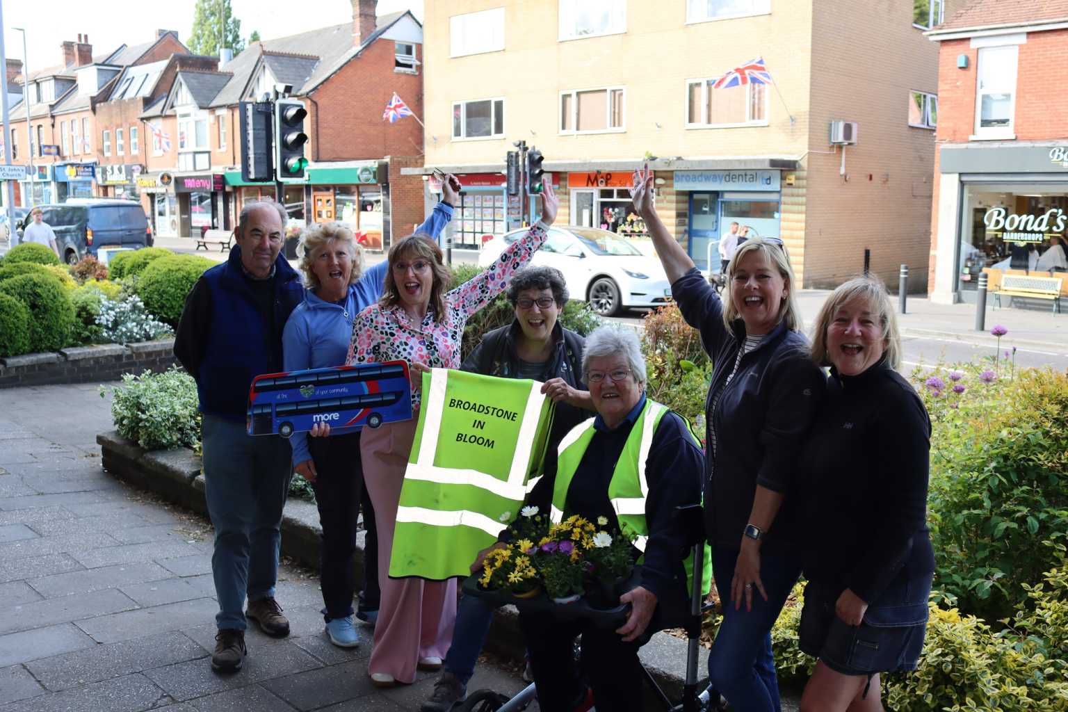 Group of people cheering in front of a flower bed holding a More bus cutout