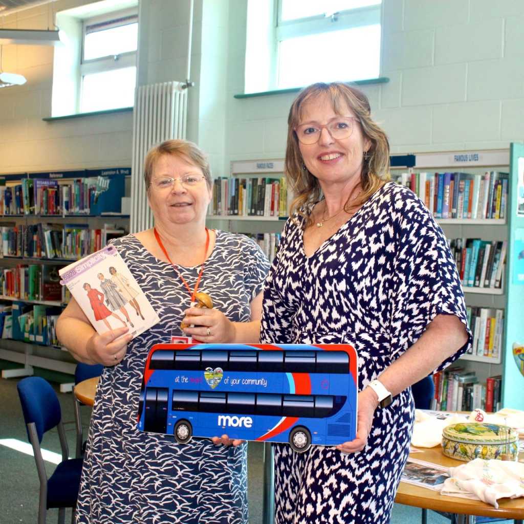 Two ladies holding a More bus cut out in a library